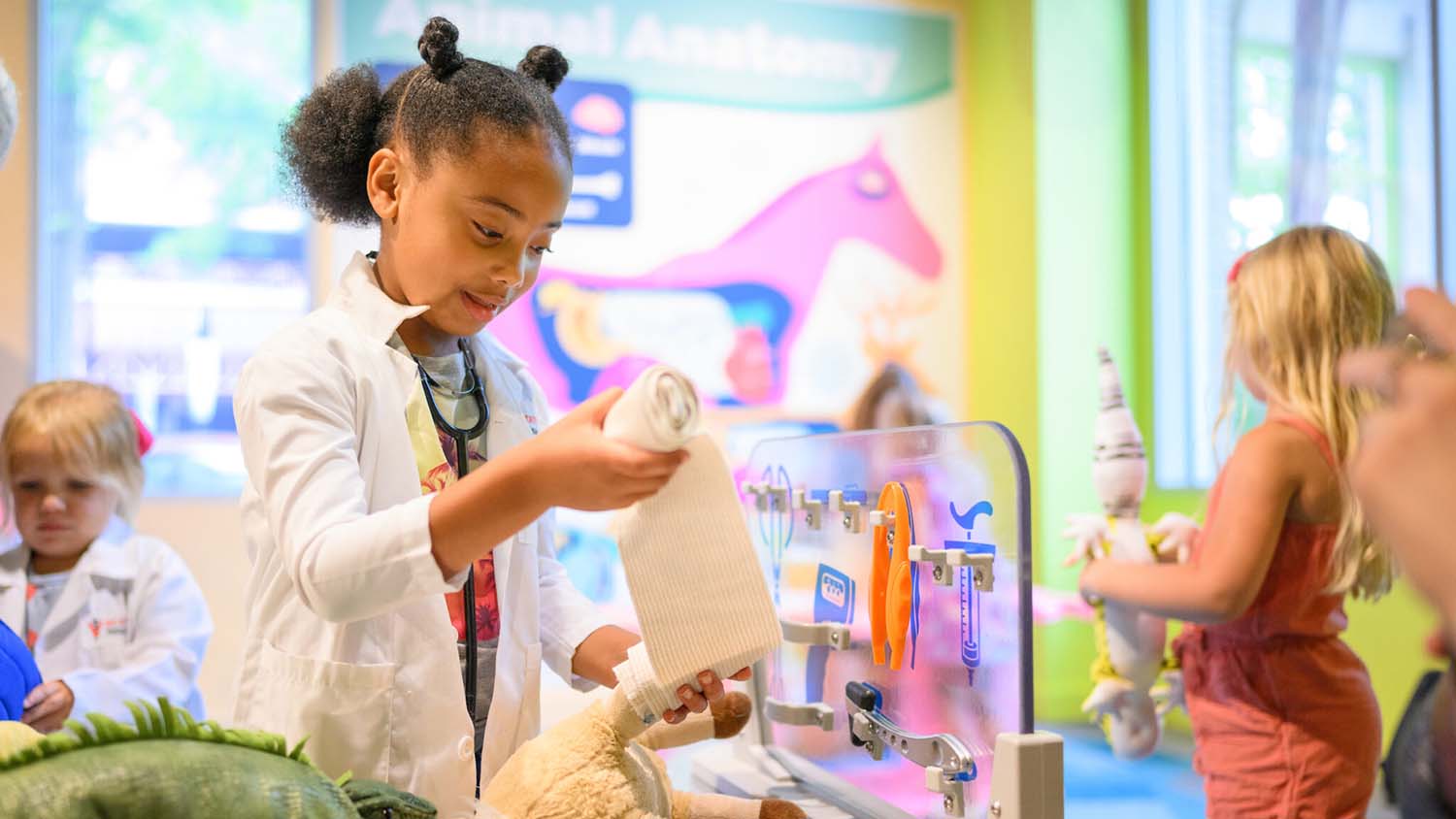 Children wear white coats and play with stuffed animals in an interactive vet-themed play area.