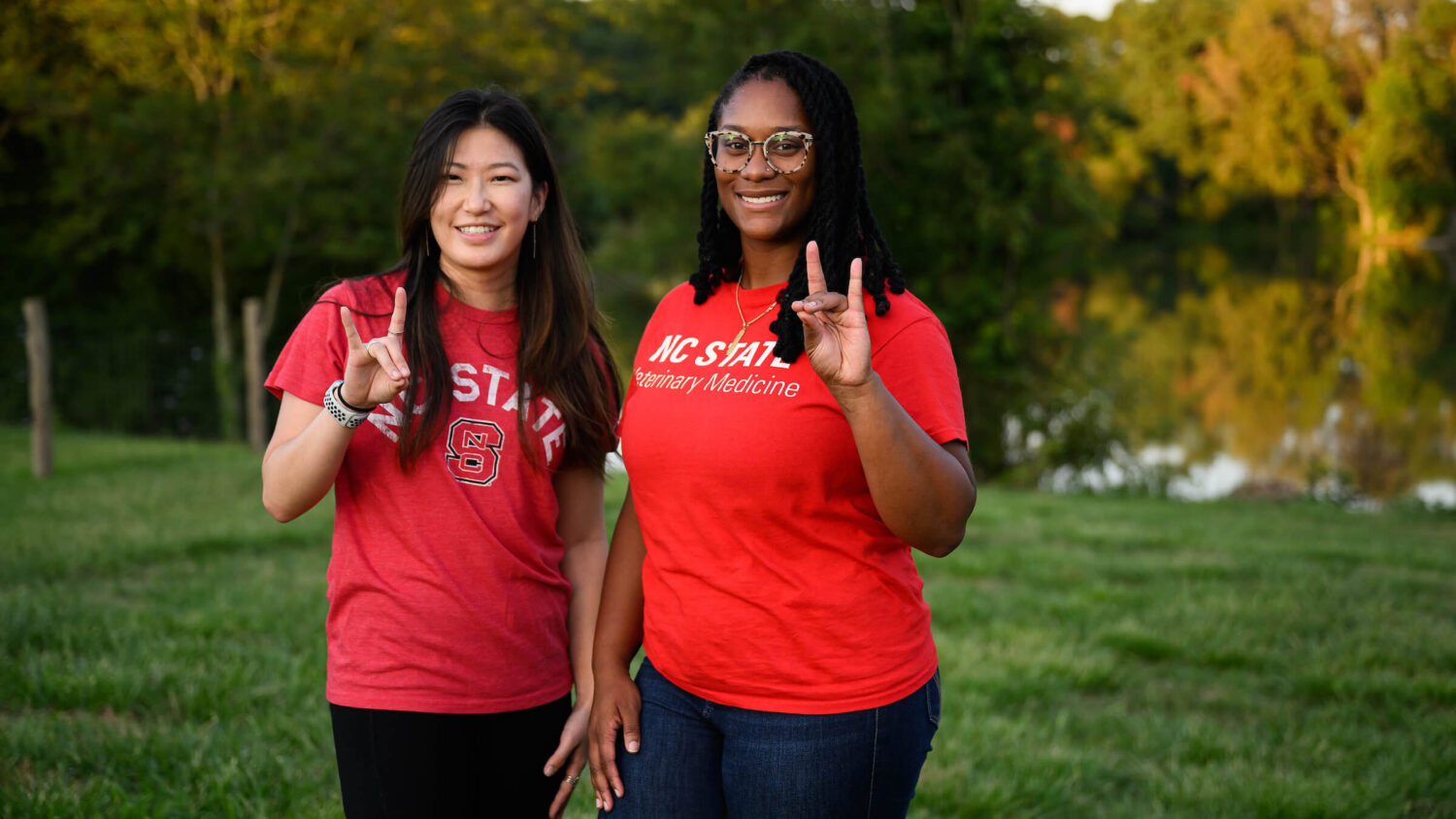 two young women pose in front of a lake while holding up the wolfpack hand sign