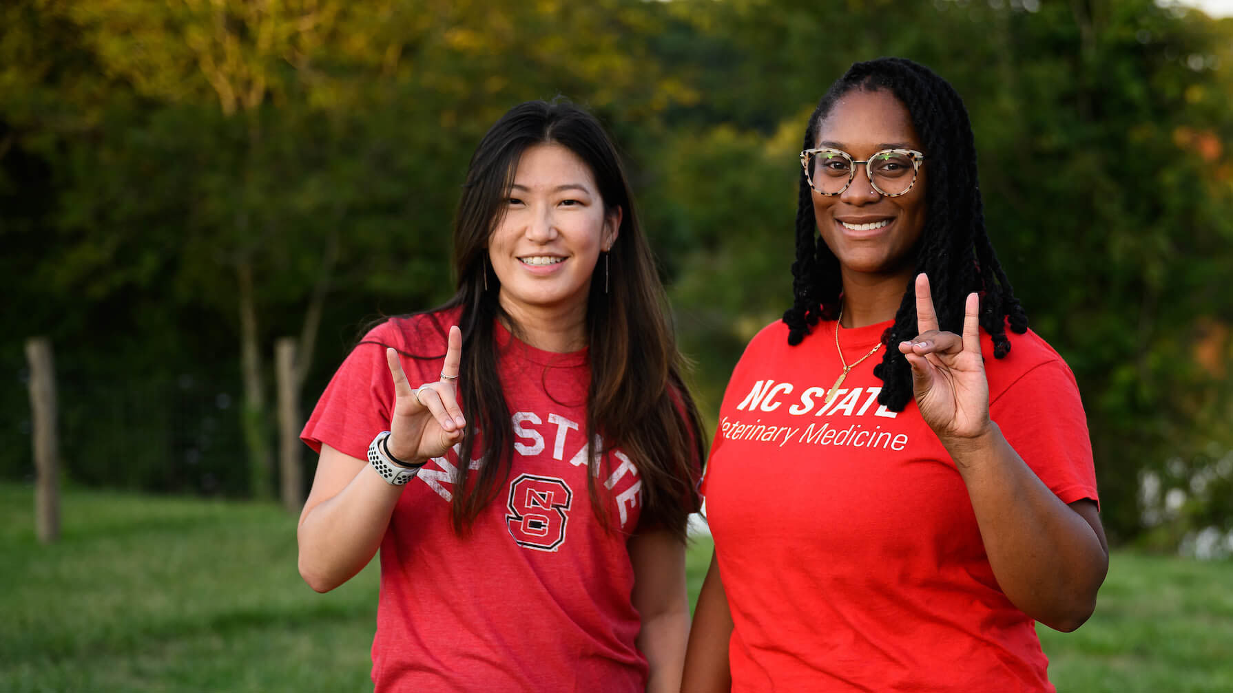 two young women pose in front of a lake while holding up the wolfpack hand sign