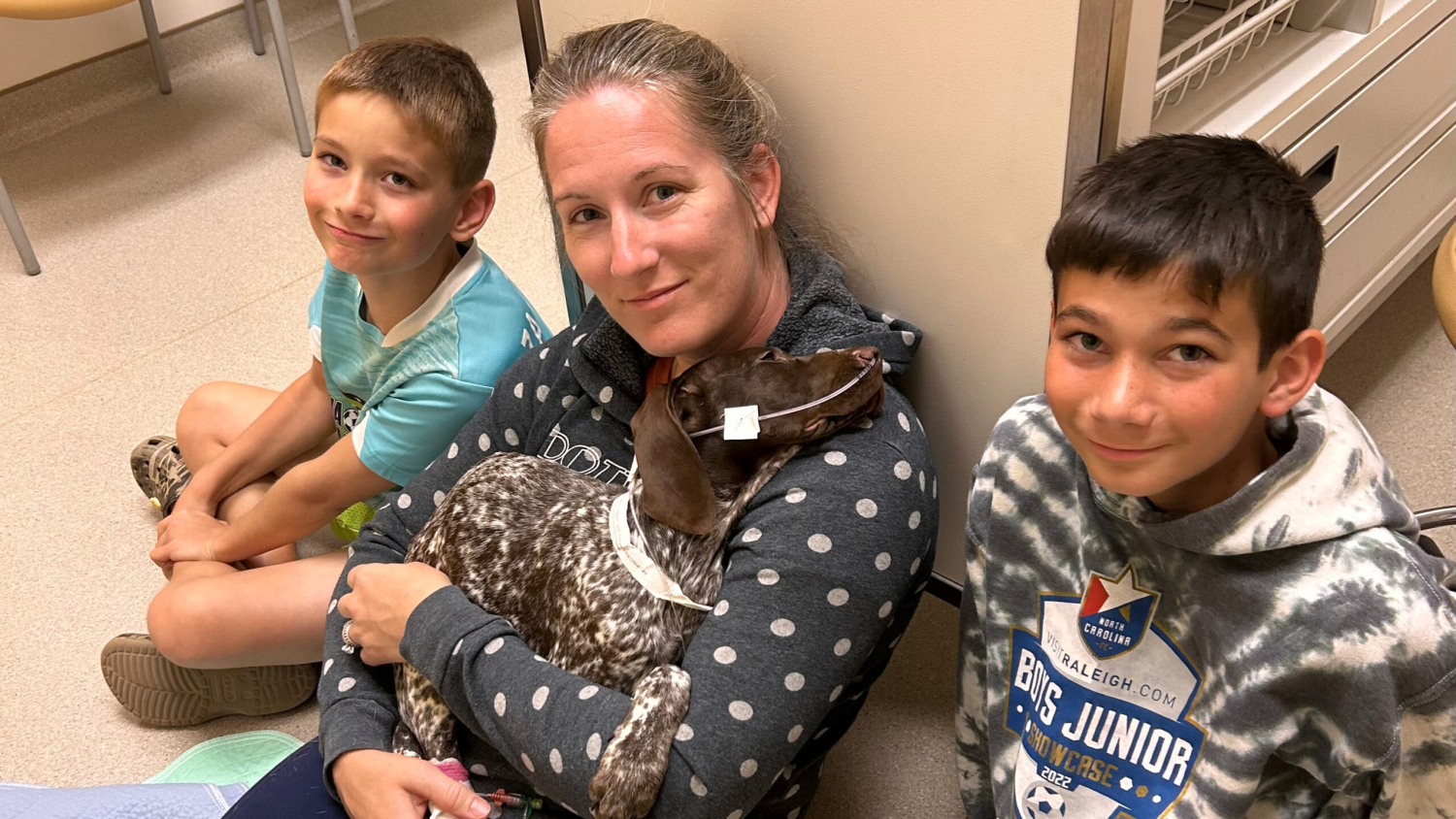A woman holding a brown-and-white speckled German shorthaired pointer puppy fitted with a nasogastric feeding tube sits between her two young sons on the floor of a veterinary hospital.
