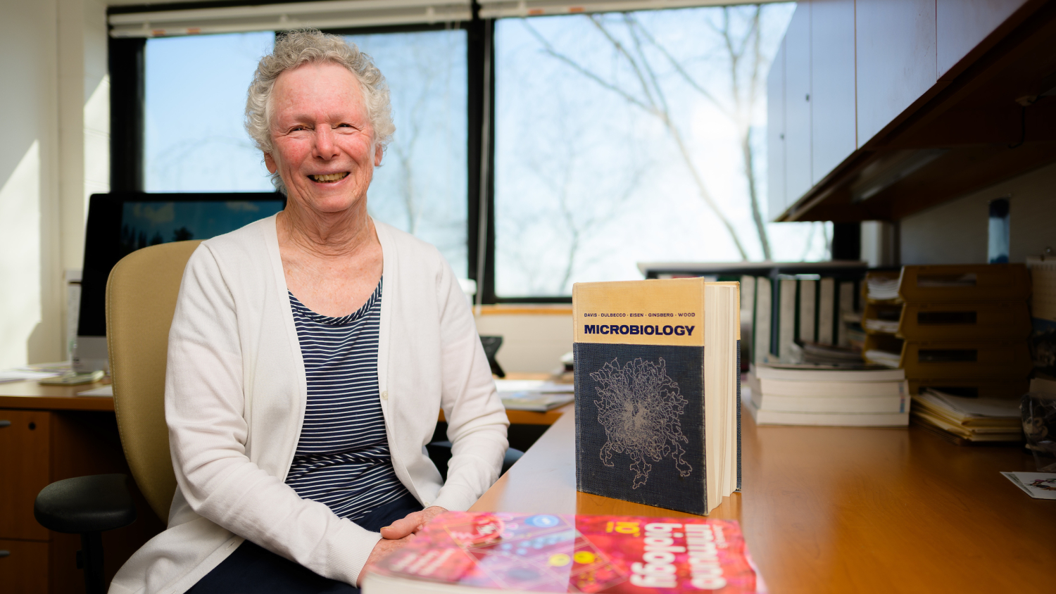 Dr. Sue Tonkonogy sits at her desk in her office. On the desk, next to her, her microbiology textbook stands on its end.