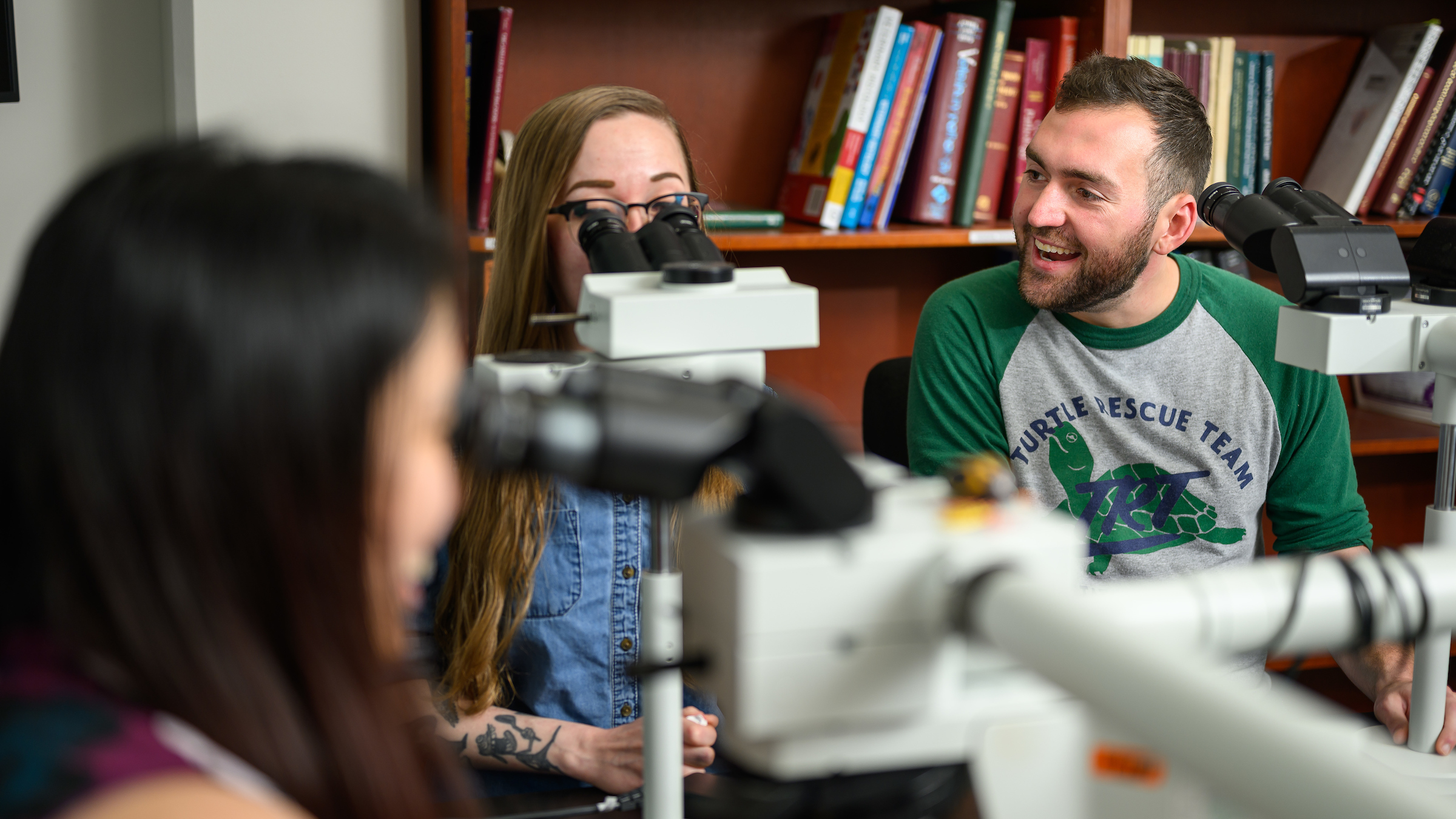 A male student smiles at two female mentors as the team sits behind a teaching microscope.