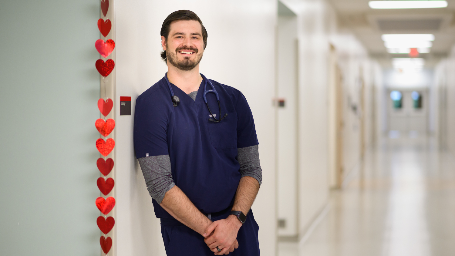 Dr. Seth Bowden, a cardiology resident at the NC State Veterinary Hospital, leans against a wall in the hallway outside the cardiology service at the NC State Veterinary Hospital. A strand of decorative hearts hangs to his right.