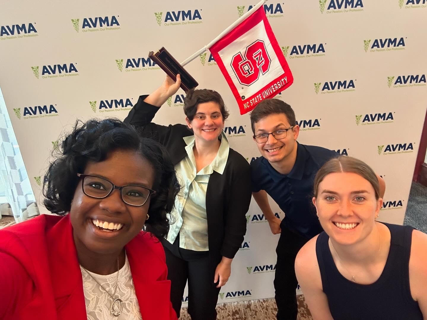 NC State SAVMA board members Anna Jones, Mary Emfinger, Max Seda and Robin Gallagher pose in front of an AVMA-branded backdrop at a conference. Emfinger, second from left, holds an NC State flag and plaque in her right hand.