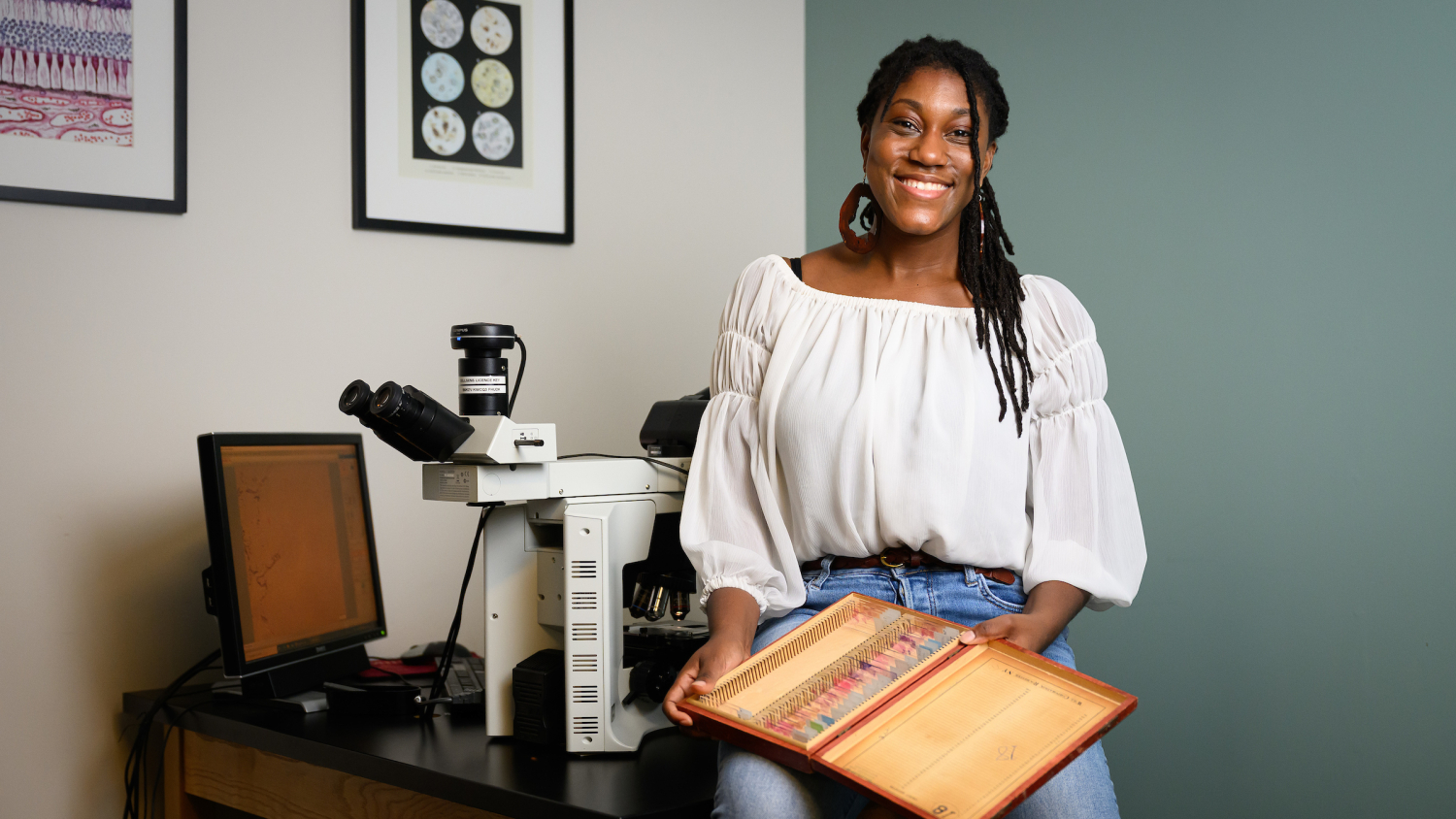 Dr. Jazz Stephens sits on the edge of a table with a box of pathology slides in her lap. Behind her, a microscope and a computer screen sit on a table to form a pathologist's work station.