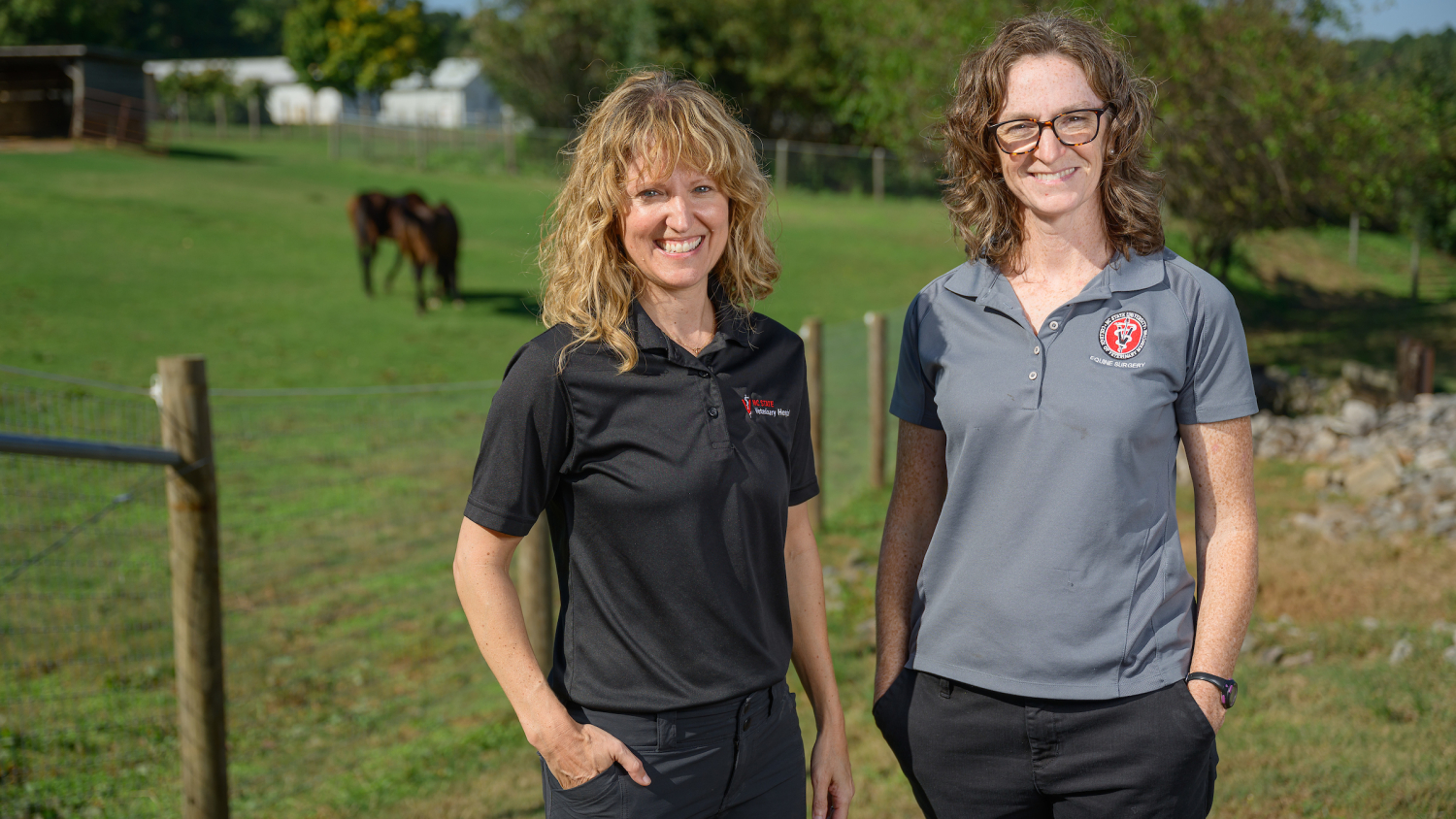 Dr. Katie Sheats, left, and Dr. Callie Fogle, two of NC State's equine health experts, pose in front of a pasture holding a grazing brown horse.