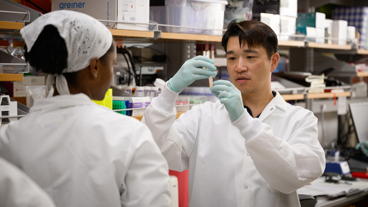 Dr. Ron Li wearing a white lab coat and pale blue gloves looks at a vial in a lab