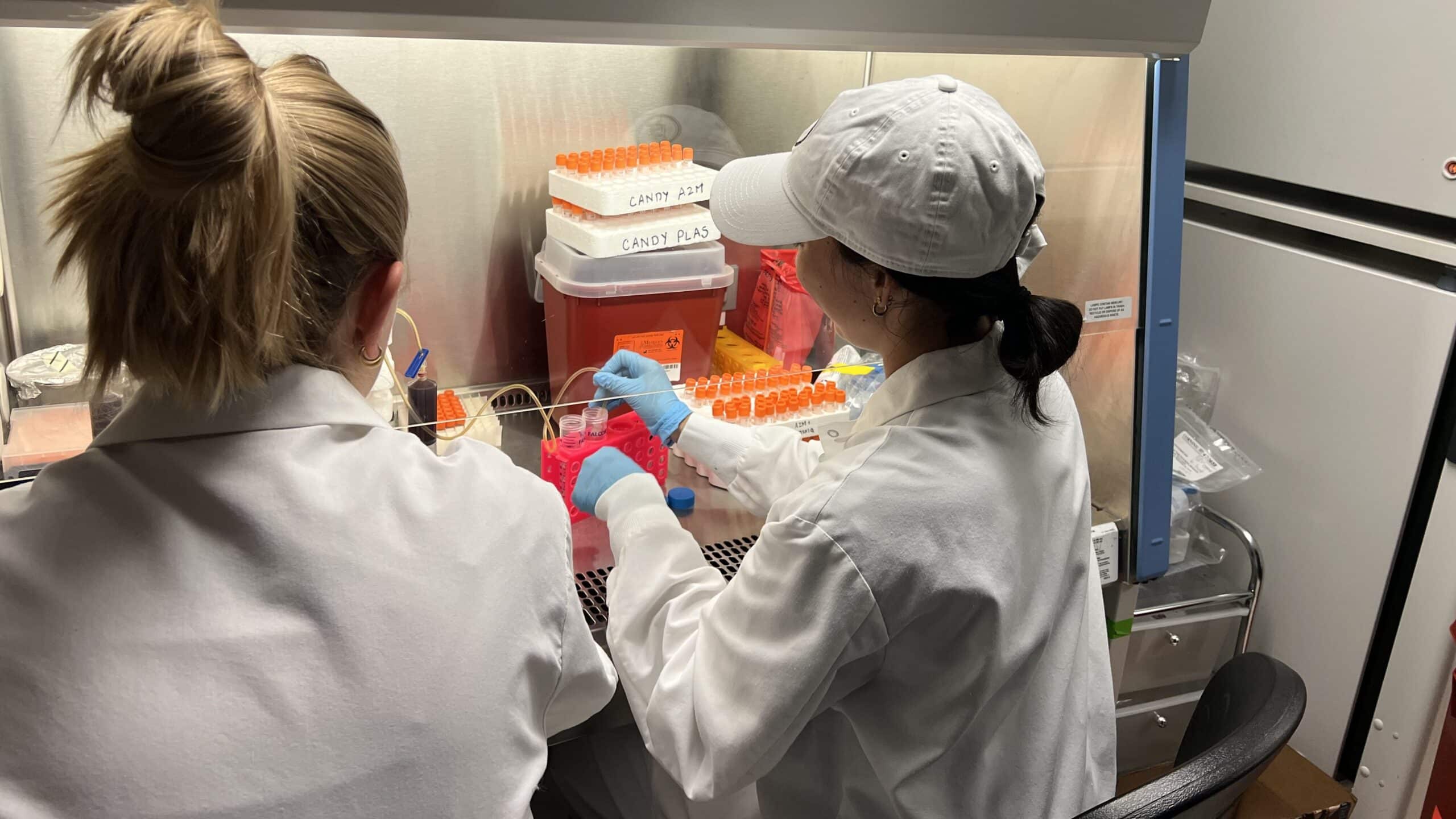 Two women in white lab coats process a blood sample at a laboratory bench.